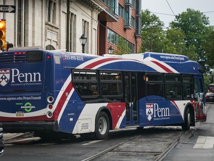 The picture is taken from the perspective of someone waiting at a stop light - at the corner ahead, a bus is seen turning onto the street. The bus has a wrapper advert covering the outside with red white and blue ribbon artwork, with a large Penn emblem and “University of Pennsylvania” decorating both the back and the side of the bus. On the back it also says “www.upenn.edu/PennTransit” and "Powered by BIODIESEL”
